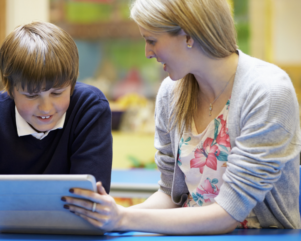 A blond-haired woman points at an i-pad with a smiling brown haired boy wearing a blue jumper
