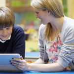 A blond-haired woman points at an i-pad with a smiling brown haired boy wearing a blue jumper